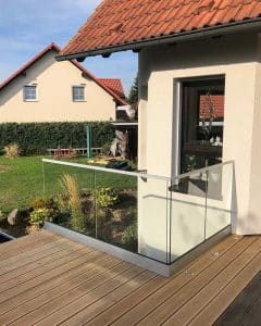 Pfostenfreies Glasgeländer auf der Terrasse Stilvolles pfostenfreies Glasgeländer mit Klarglas und silbernem Handlauf, montiert auf Holzterrasse am weiß verputzten Haus mit Ziegeldach.