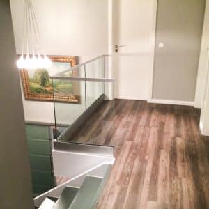 Glasgeländer im Treppenbereich mit weißem Handlauf Pfostenfreies Glasgeländer im oberen Treppenbereich mit weißem Handlauf und Klarglas, montiert auf dunklem Holzboden in modernem Innenraum.