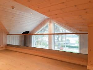 Innen montiertes Glasgeländer vor Fensterfront Pfostenfreies Glasgeländer im Innenbereich vor großem Dreiecksfenster, mit Holzverkleidung an Decke und Wänden und Ausblick ins Grüne.