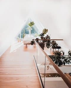 Elegante Innenraumlösung mit Glasgeländer Helle und stilvolle Dachwohnung mit pfostenfreiem Glasgeländer und Holzhandlauf, eingerichtet mit Pflanzen, Holzfußboden und Bett am großen Dreiecksfenster.