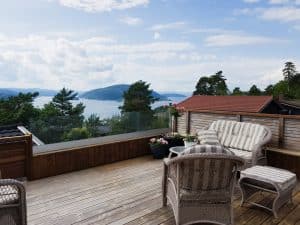 Pfostenfreies Glasgeländer auf Terrasse mit Aussicht Stilvolles pfostenfreies Glasgeländer auf holzverkleideter Terrasse mit Blick auf Wald und Wasser, ergänzt durch Lounge-Gruppe und Blumentöpfe.
